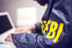 foto de um computador (fora de foco) sendo operado por uma pessoa de jaleco azul marinho, com a inscrição “FBI” na manga.