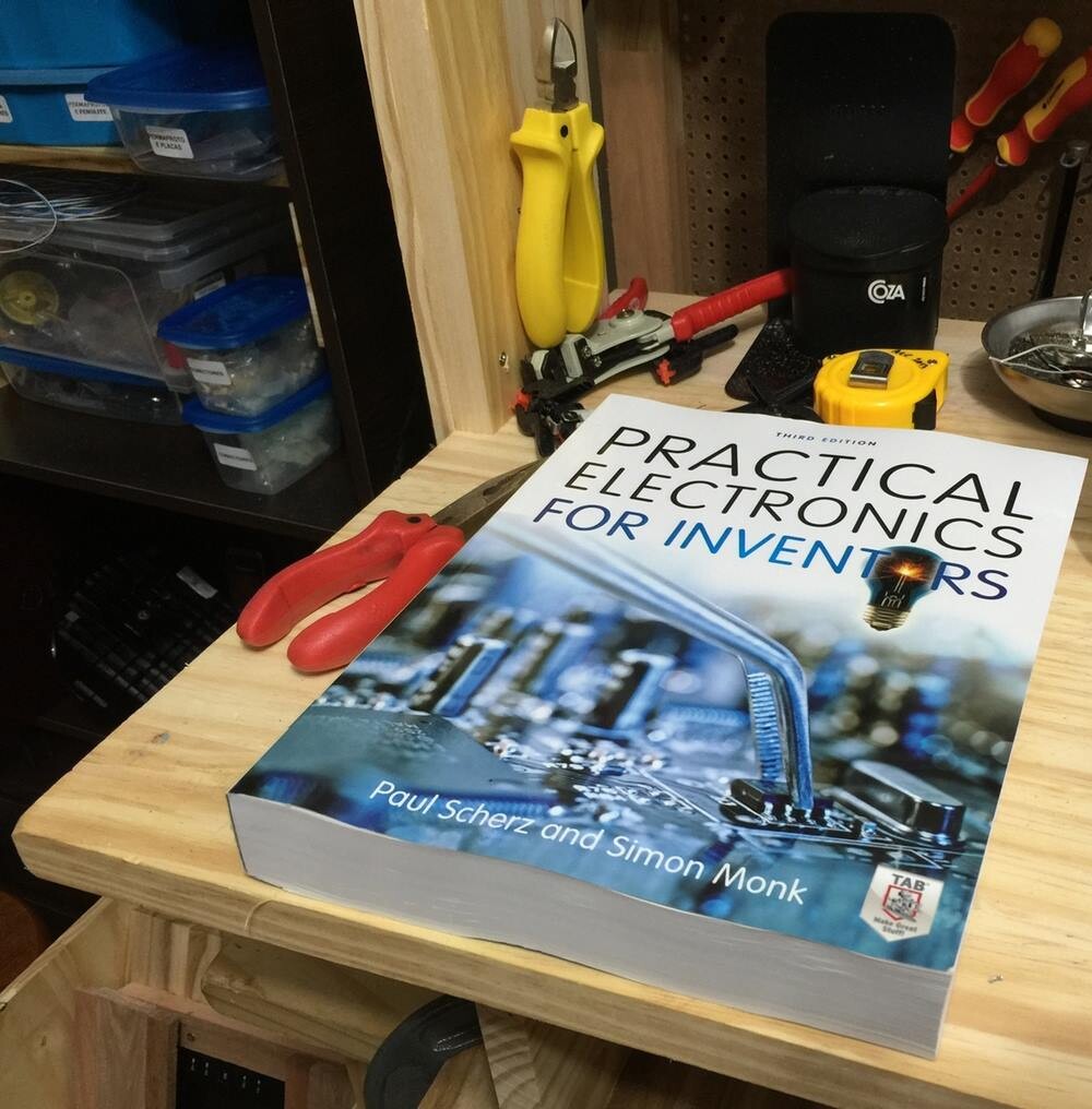 Exemplar do livro Practical Electronics for Inventors, sobre a minha bancada de madeira, cercado de ferramentas