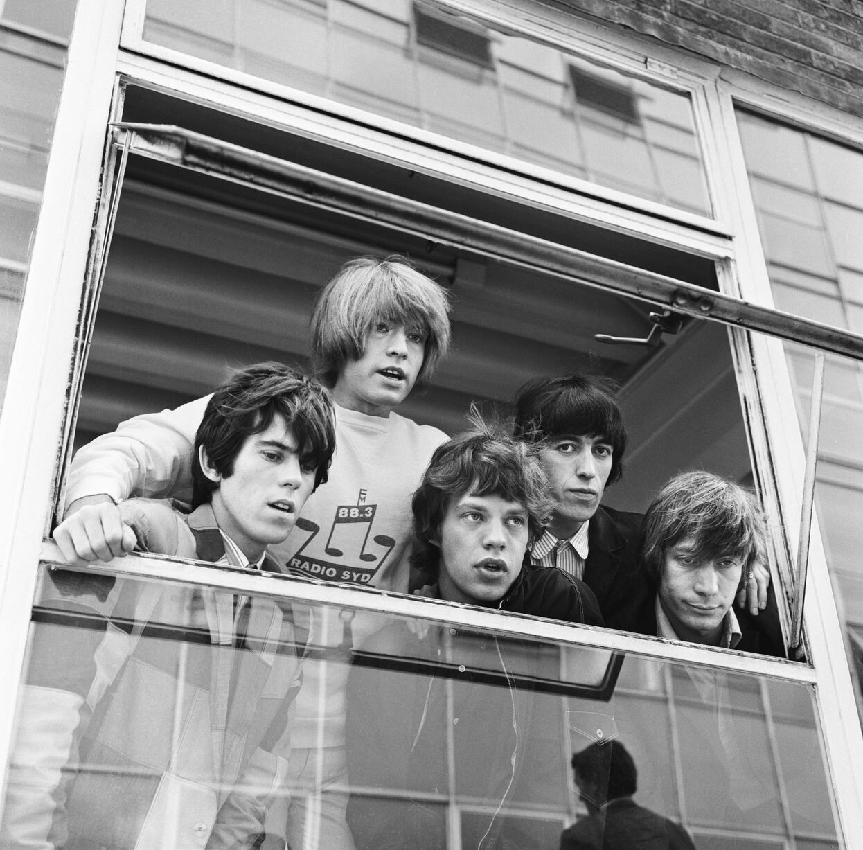 Foto em preto e branco dos Rolling Stones em 1965, olhando por uma janela.