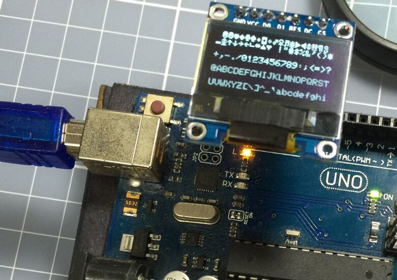 Placa Arduino Uno com um display OLED pouco maior que uma moeda de um real conectado a ela, exibindo as letras do alfabeto na tela