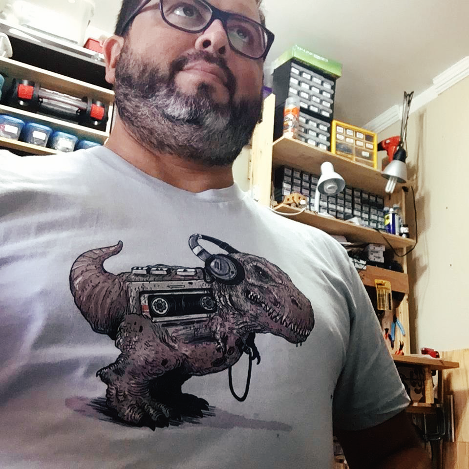 Foto minha com duas bancadas ao fundo e com camiseta cuja estampa é um dinossauro ouvindo um walkman.