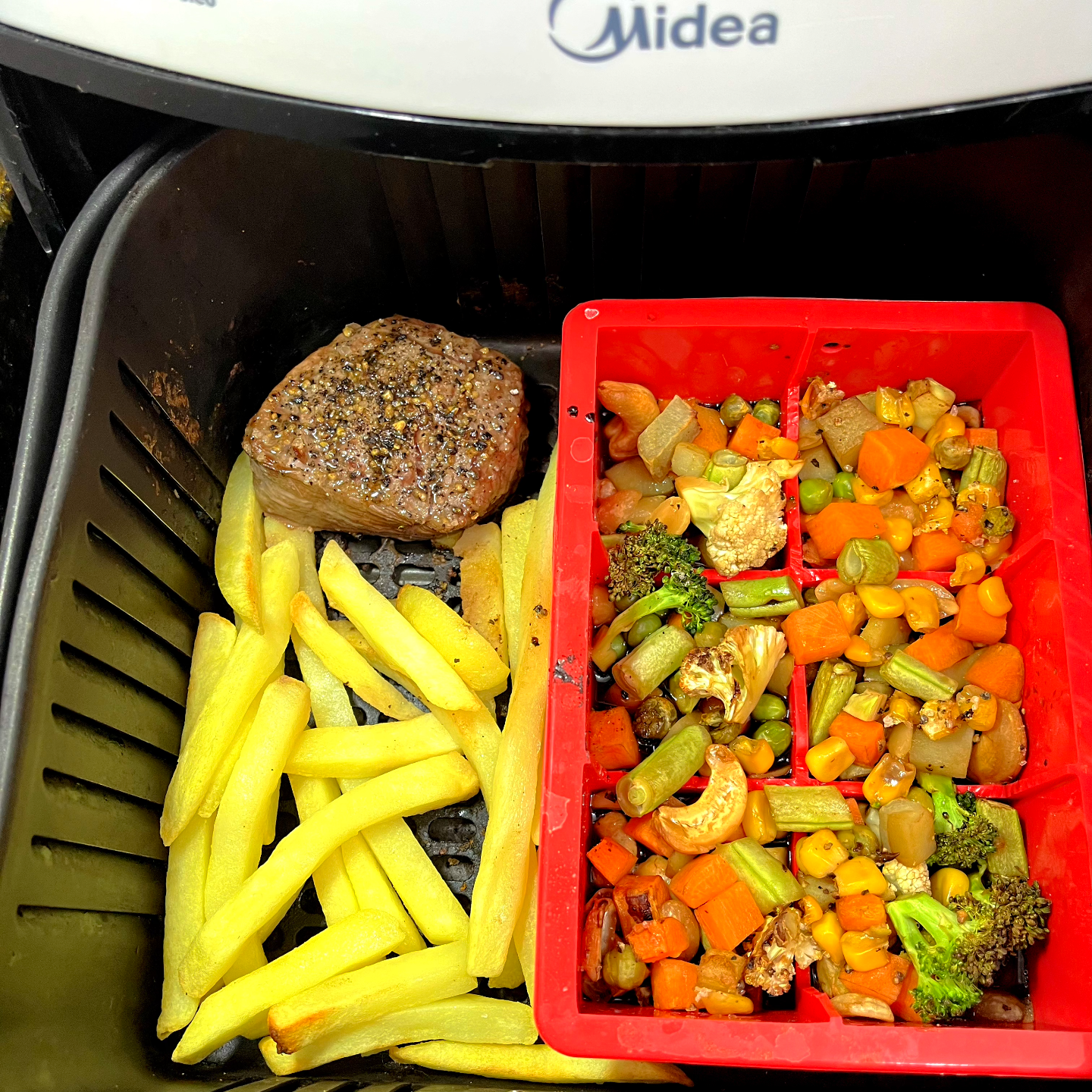 Airfryer aberta mostrando um medalhão de carne coberto de pimenta do reíno moída em casa, 26 palitos de batata frita, e uma forma de silicone repleta de legumes picados e shoyo