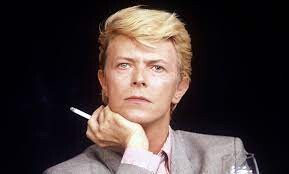 David Bowie, cigarro em punho, de traje social, encara a câmera