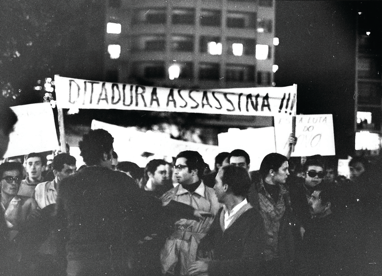 Foto em preto e branco de pessoas em uma manifestação, com uma faixa ao fundo onde se lê: DITADURA ASSASSINA