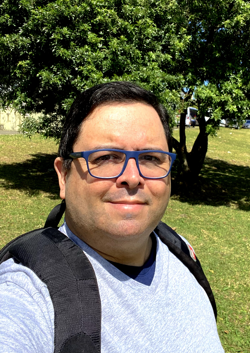 Homem de mochila, casaco e óculos com armação azul, olhando para a câmera, com gramado e árvore ao fundo