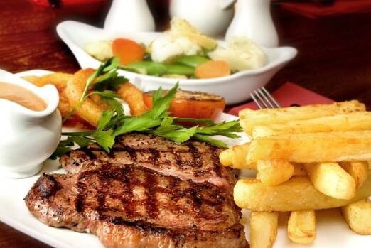 Um prato de bife, com batatas fritas cenográficas, aneis de cebola, jarro de molho e uma salada ao fundo