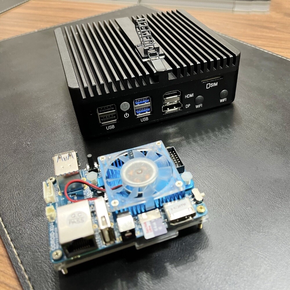 Ao fundo, em foco, um servidor estilo “PC industrial“, pequeno, num gabinete preto, com portas USB e HDMI visíveis. <br><br>À frente, ligeiramente fora de foco, uma placa Odroid XU4.