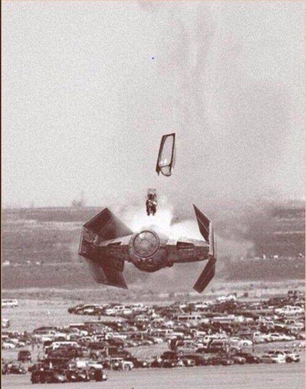 Foto em preto e branco mostra um piloto ejetando de um tie fighter (dos filmes Star Wars), a baixa altitude, sobre um estacionamento de carros