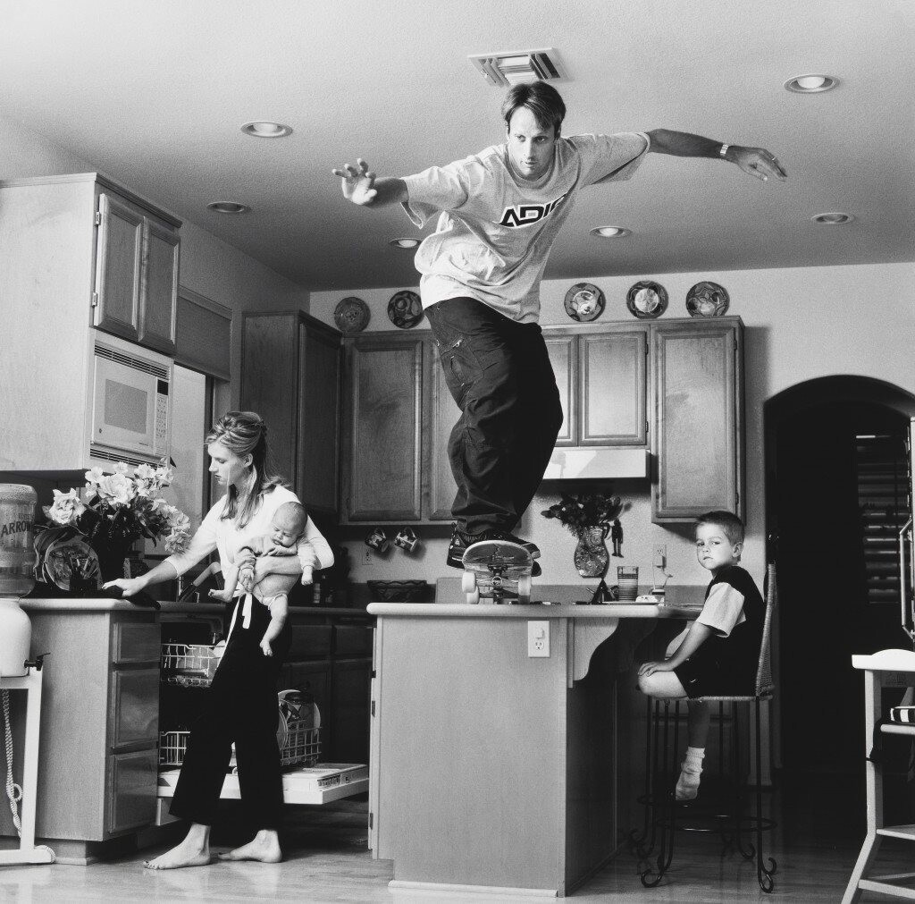 Foto em preto e branco de homem jovem se preparando para dropar de skate de cima do balcão da cozinha, sendo ativamente ignorado por mulher jovem que segura um bebê, enquanto uma criança pouco mais velha olha para a câmera expressando desprezo pela situação