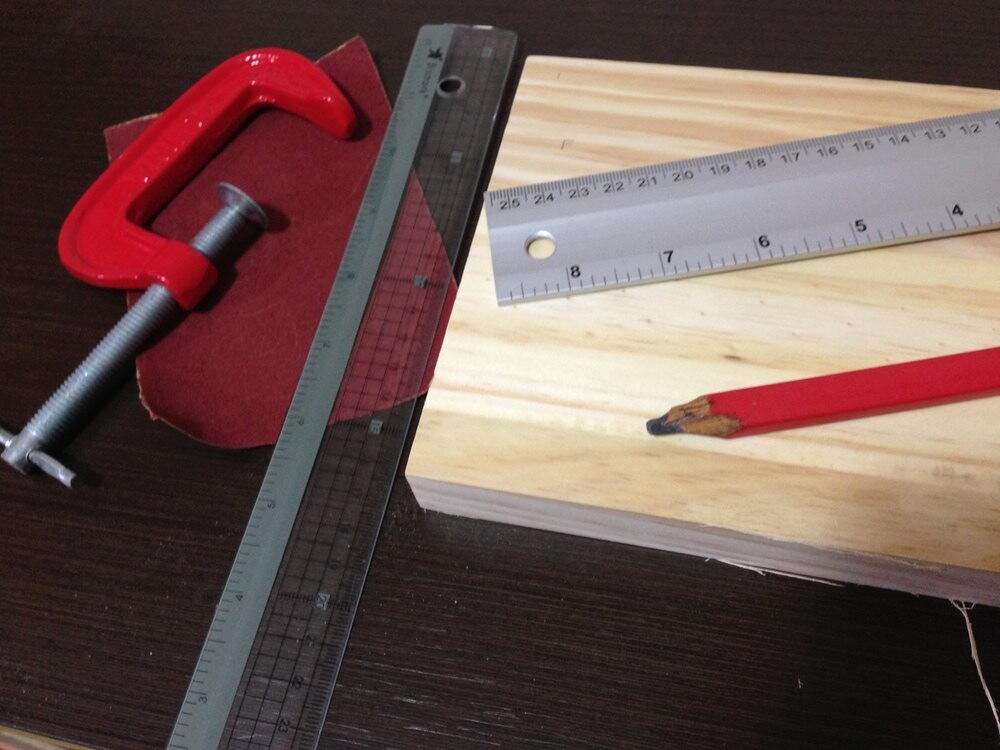 Uma tabua de pinus, duas réguas, um toco de lápis, lixa e um grampo-sargento parcialmente aberto