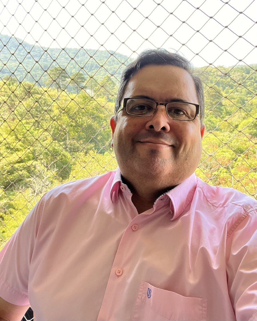 Selfie minha olhando pra câmera, sorrindo, de camisa rosa de manga curta e com detalhes em azul, tendo a área de preservação permanente ao fundo