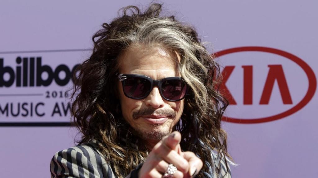 Steven Tyler, de óculos escuros,  aponta para a câmera