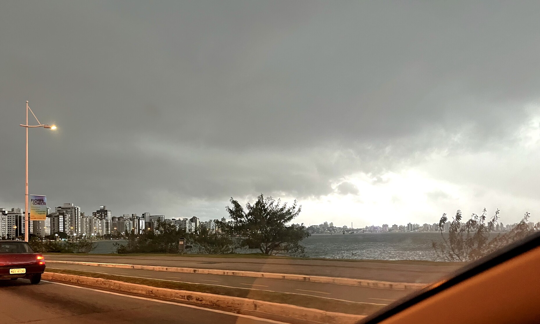 Vista de dentro do carro, mostrando a ciclovia da beira-mar norte, trecho da baía e o céu plúmbeo