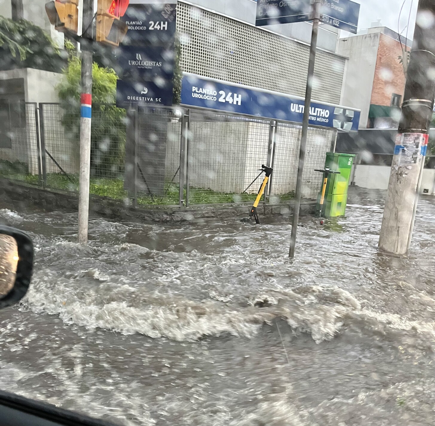 Vista de dentro do carro, mostrando a calçada, a sarjeta e a pista tomadas pela água acumulada, e um patinete motorizado mal posicionado para lidar com a situação 