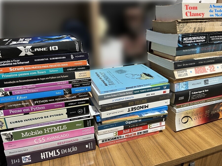 3 pilhas de livros, sendo que a primeira é mais com temas de TI (especialmente web, python e git), a do meio são coisas de gestão e assemelhados, e a da direita é literatura mais variada, incluindo temas de História do Brasil, neuro, romances e mais.