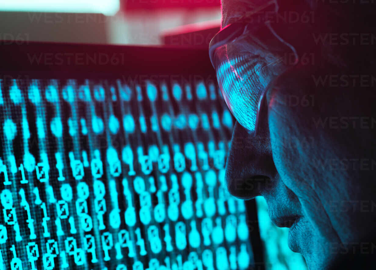 Homem de óculos escuros, no escuro, observa uma tela repleta de dígitos binários