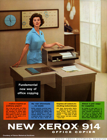 Anúncio da Xerox 914, com uma jovem trajada de azul, mesma cor do relógio na parede. Ela está ao lado da volumosa e inovadora copiadora.