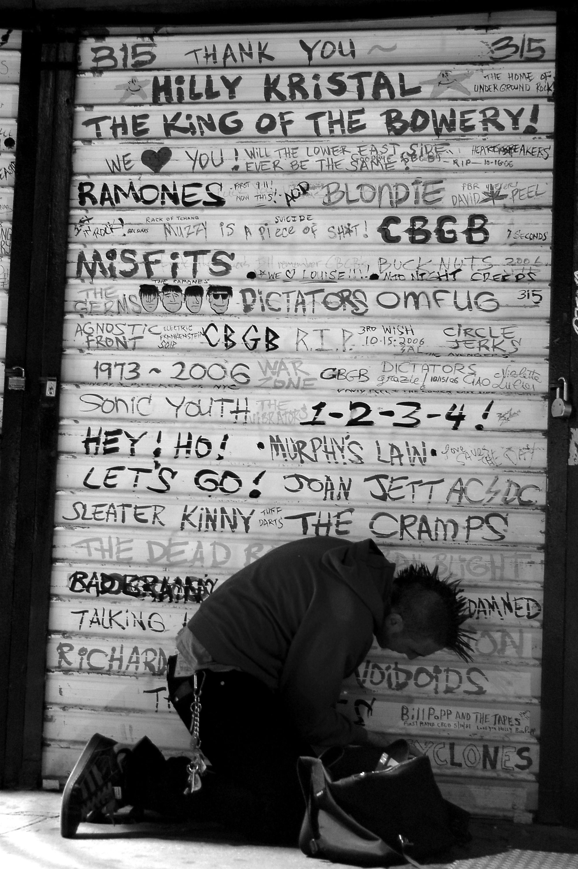 Um punk ajoelhado junto à porta fechada do CBGB, decorada com os nomes de várias das bandas e artistas que neles se apresentaram entre os anos 70 e o início do século XXI