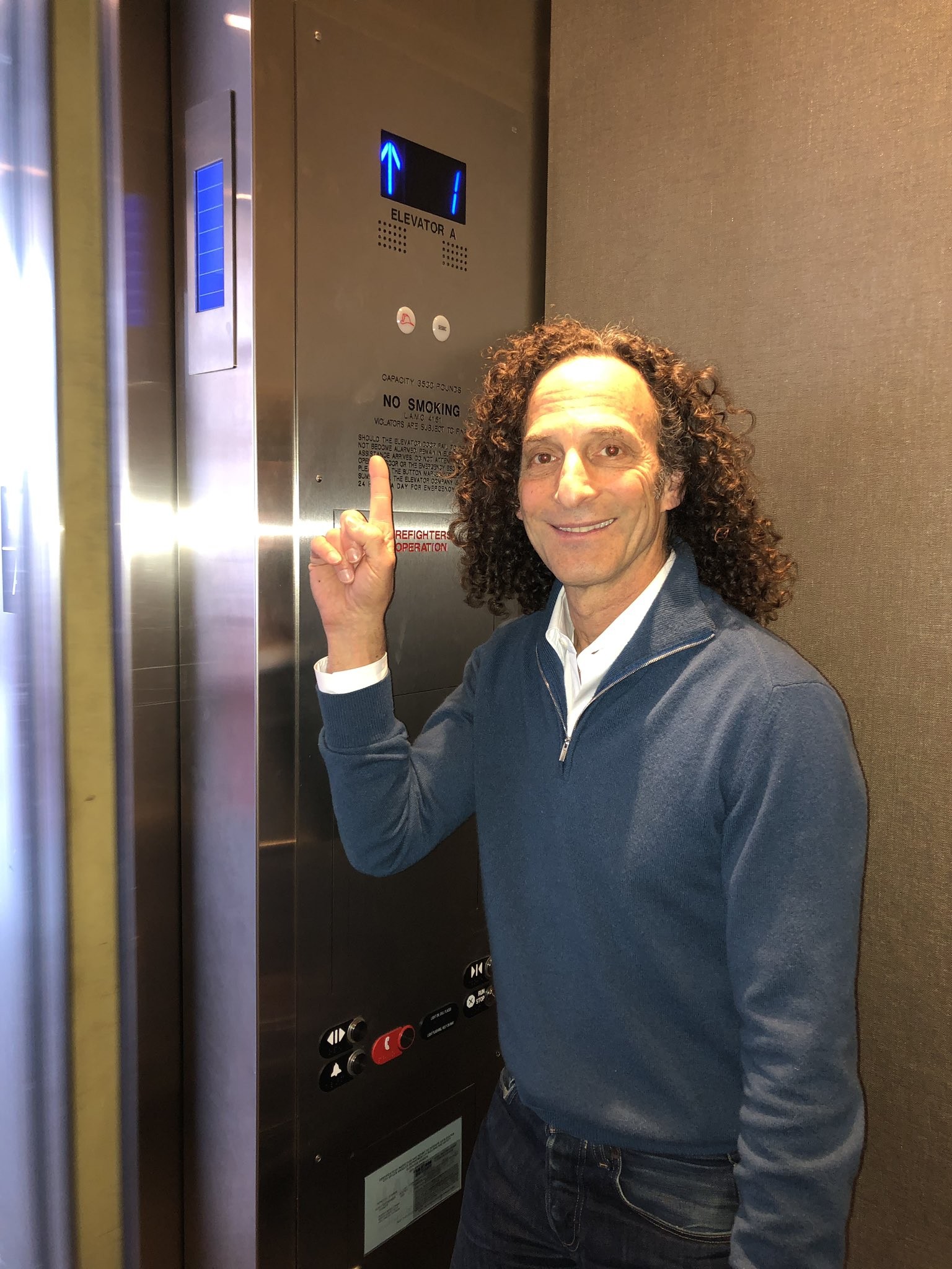 Kenny G, no elevador, sorri para a câmera e aponta para cima