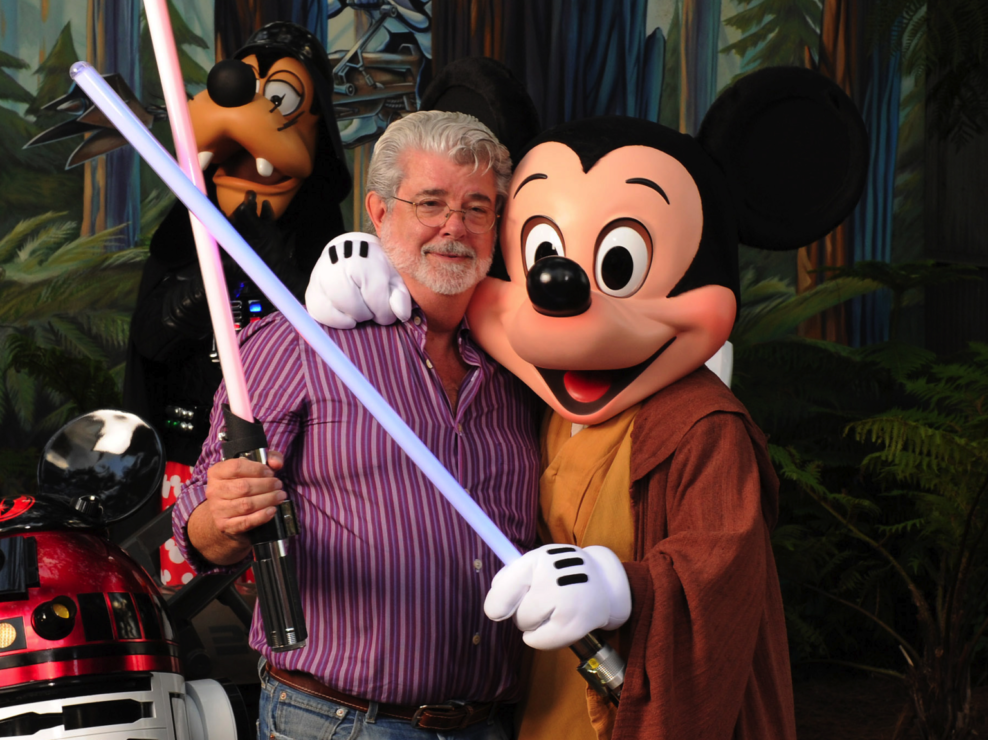 George Lucas segura um sabre de luz de brinquedo, abraçado ao Mickey com um robe jedi