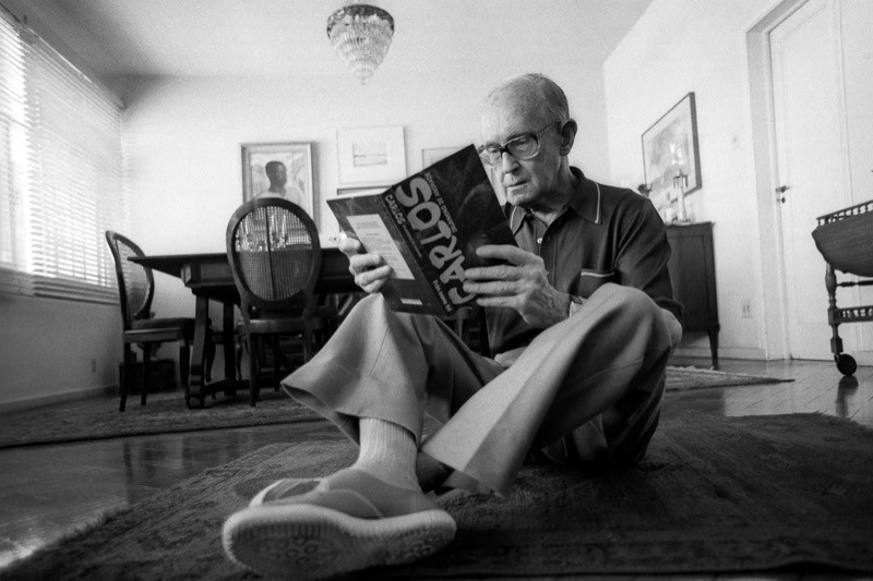 Drummond posa para a fotografia sentado no chão de seu apartamento, lendo uma antologia de sua obra