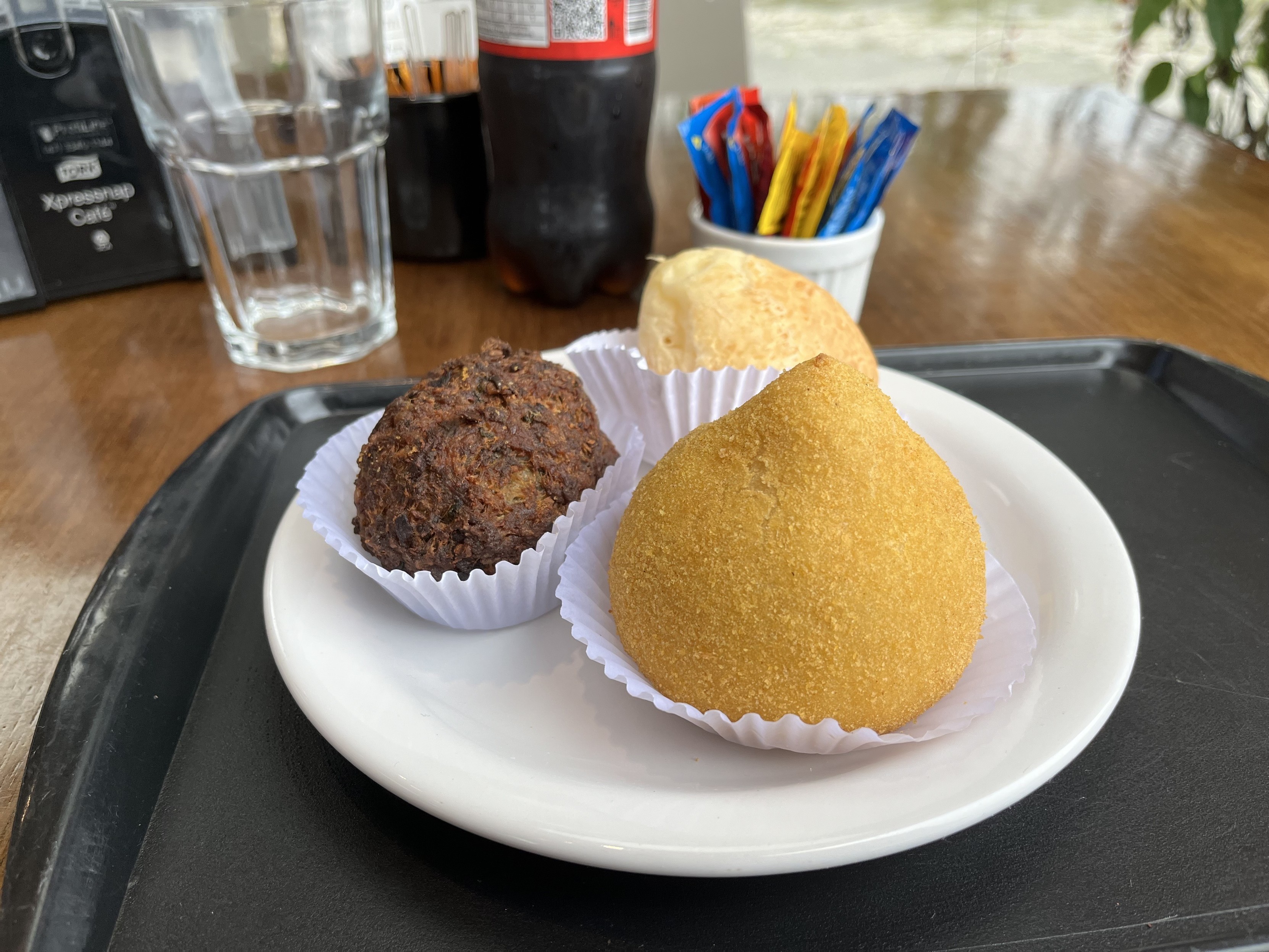 Prato com uma coxinha, uma almôndega e um pão de queijo. Ao fundo, uma coca-cola