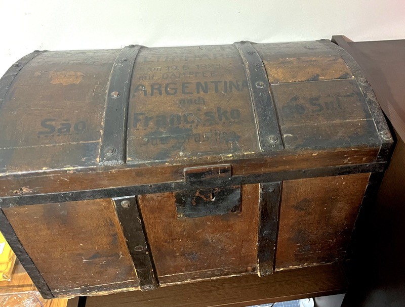 Baú de madeira com reforços em metal, em cor escura, com inscrições na tampa indicando o nome da família, a data de partida (19/6/1924), o destino do navio (Argentina) e o porto de parada (São Francisco do Sul)