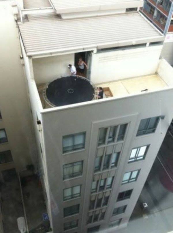 3 adultos instalando uma grande cama elástica junto ao parapeito no terraço acima do 5º andar de um prédio
