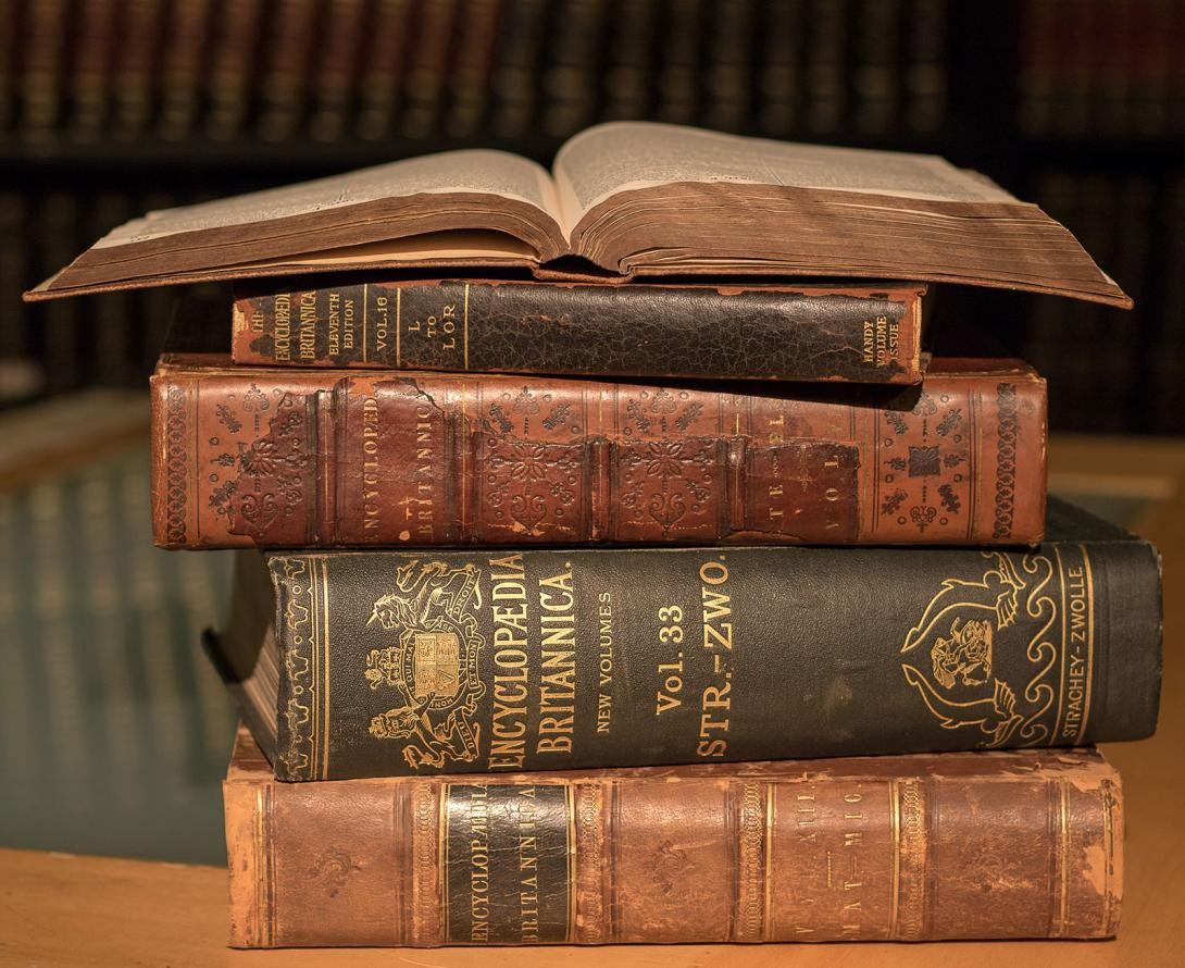 Cinco volumes antigos da Britânica, empilhados, e com um deles aberto