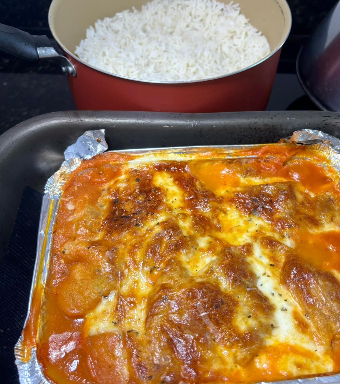 Um filé à parmegiana, ainda na travessa de aluminio em que foi assado, ao lado de uma panela de arroz branco