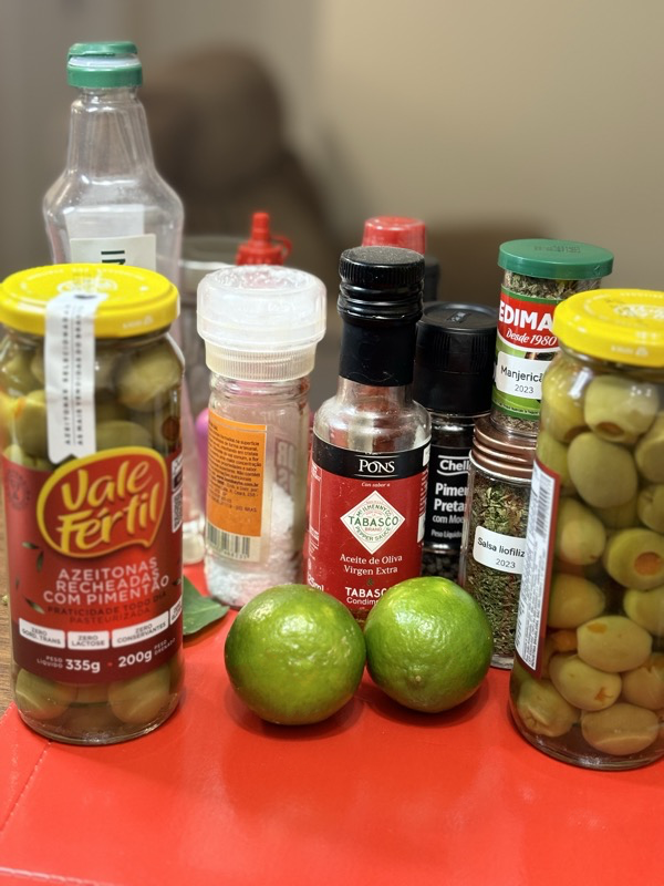 Ingredientes espalhados sobre a bancada: 2 vidros de azeitonas em conserva, 2 limões, manjericão, salsa, azeite apimentado, moinho de sal e mais
