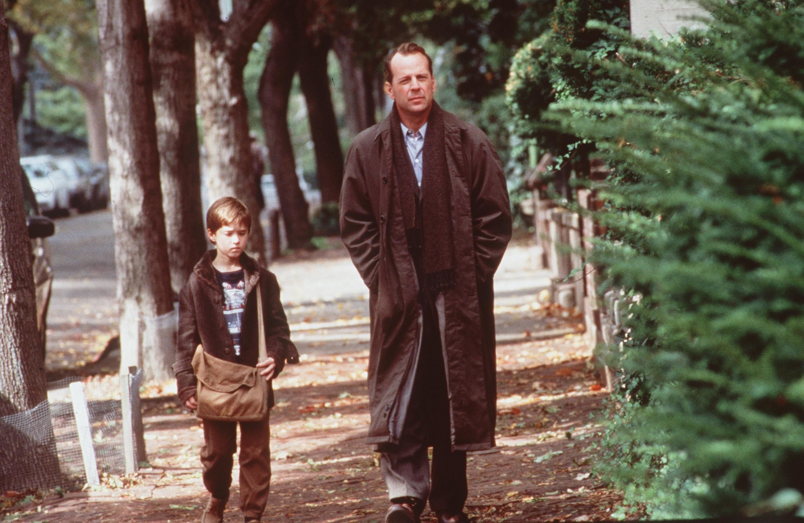 Ambos de frente para a câmera, o protagonista infantil de um dos grandes plot twists de Hollywood caminha por uma rua arborizada, com uma bolsa a tiracolo, ao lado do personagem que pensa ser seu psicólogo, vivido por Bruce Willis.