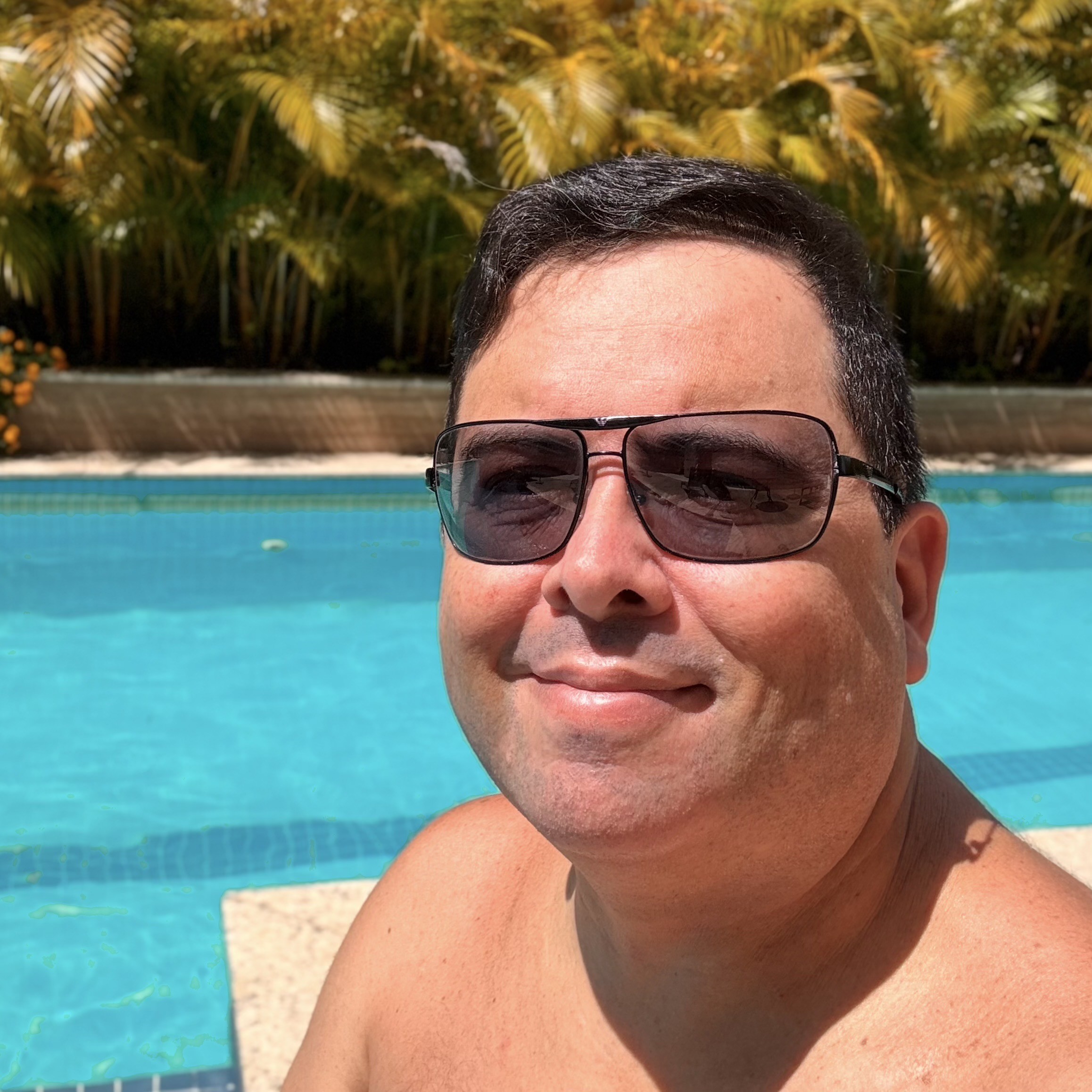 Foto minha, sem camisa, ao sol, olhando para a câmera, de óculos escuros, no ambiente de uma piscina