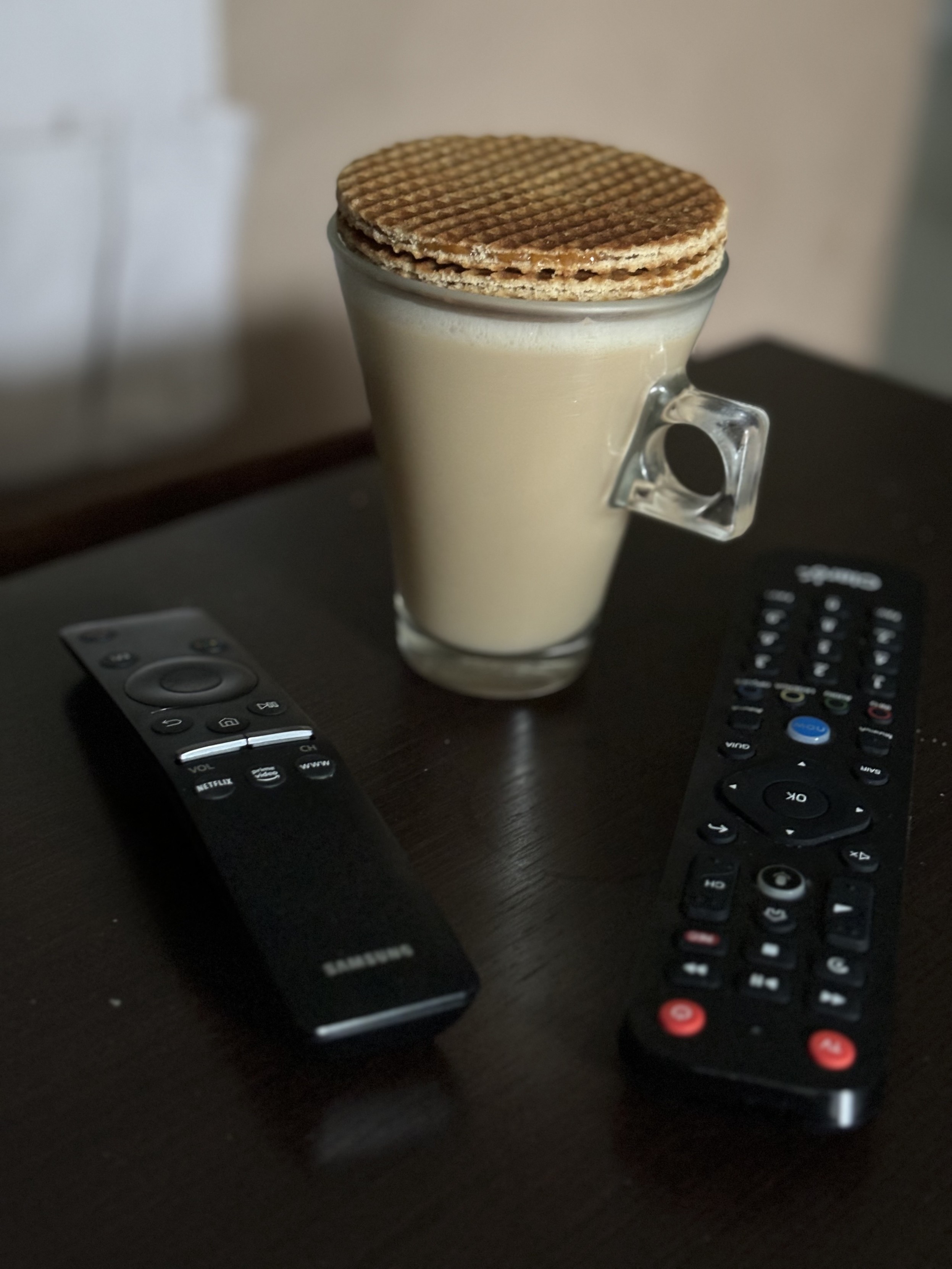 Foto da minha xícara de café com leite, entre 2 controles remotos e tampada por 2 biscoitos tipo stroopwafel 