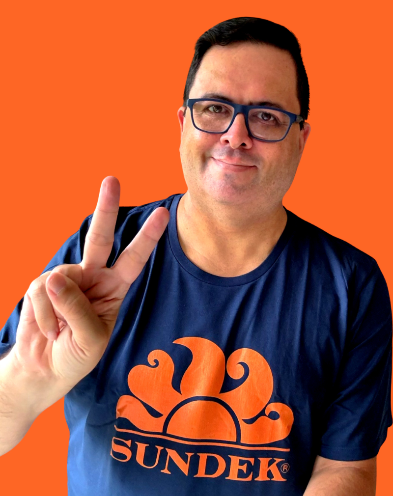 Eu, de cabelo curto, mostrando 2 dedos para a câmera num sinal de vitória, usando uma camiseta azul com estampa da Sundek em cor laranja