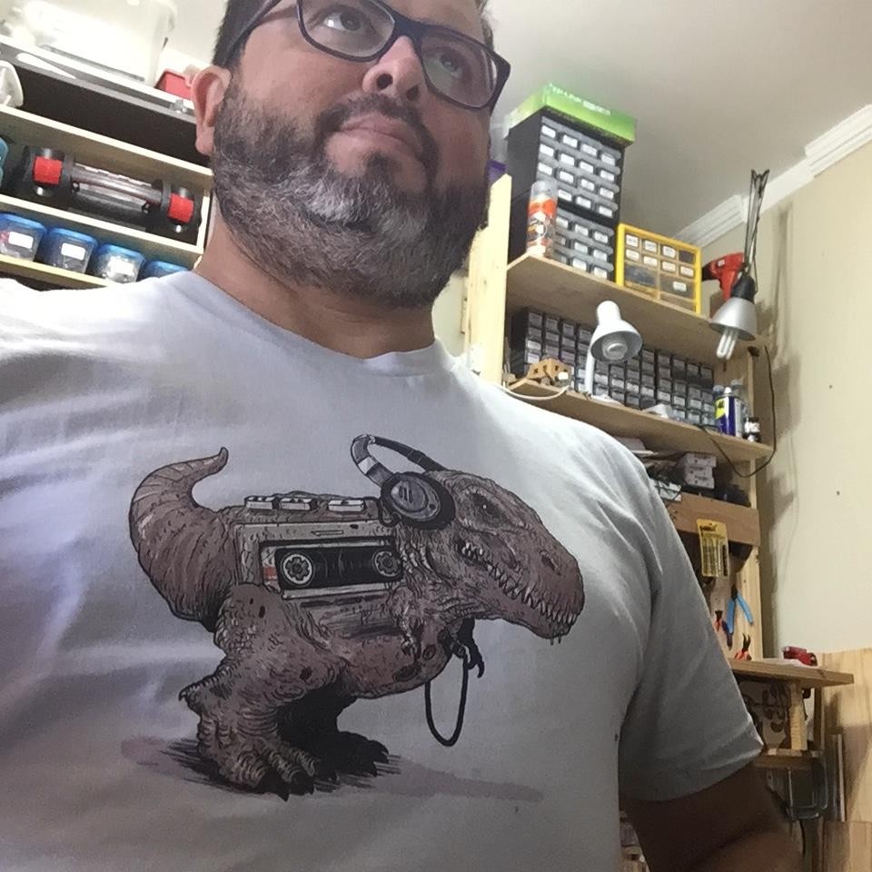 Selfie minha, com a bancada do laboratório de casa ao fundo, ainda com barba, e usando uma camiseta com estampa de um tiranossauro ouvindo walkman