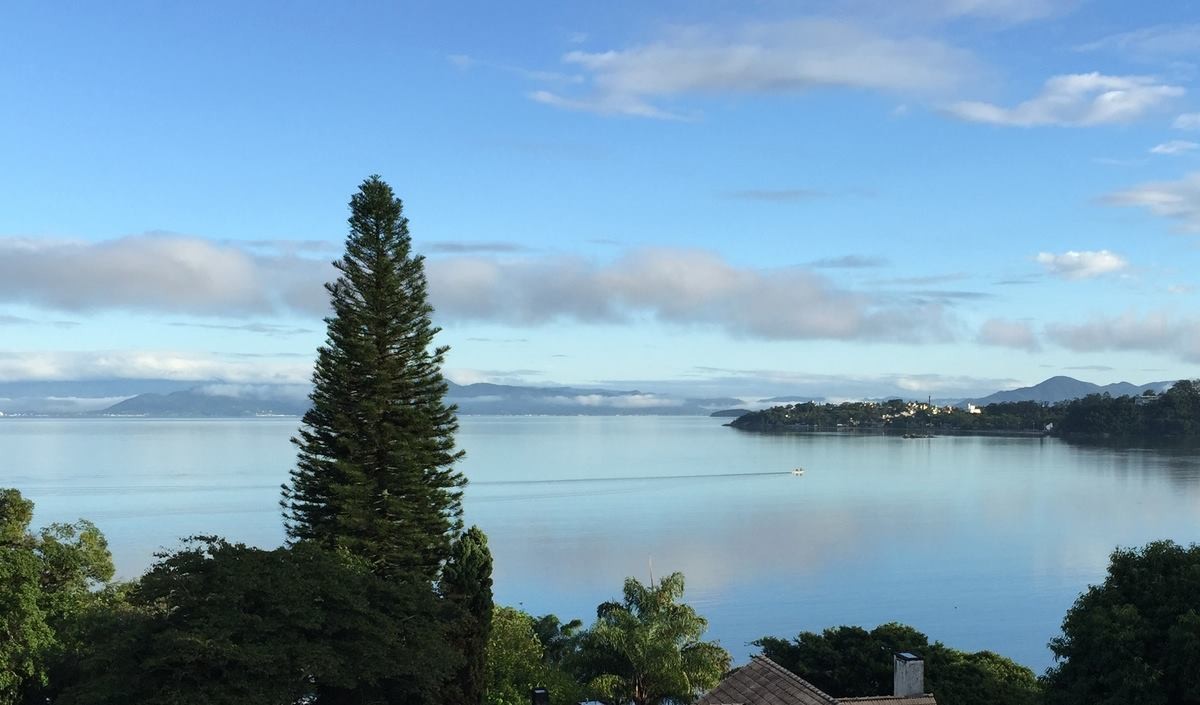 Foto da baía norte em Florianópolis, vista do bairro João Paulo, com um barco de pescadores marcando a superfície lisa, o céu de outono, e ao fundo as nuvens na serra
