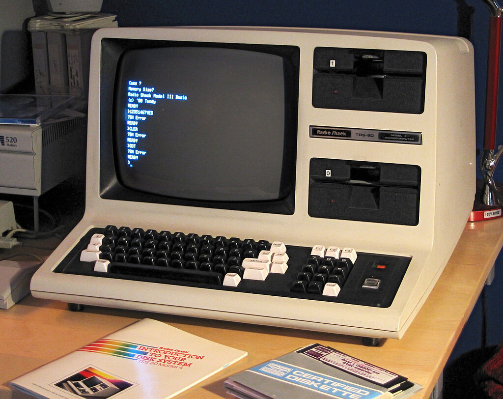 Um TRS-80 Modelo IV, ligado, com 2 manuais sobre a mesa