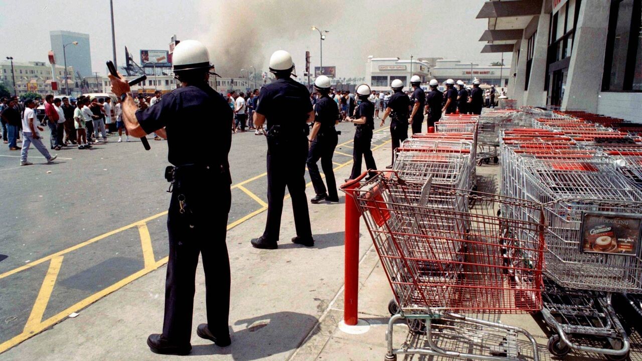 Policiais enfileirados na calçada, com multidão na rua em frente a eles, e fumaça de incêndios ao fundo