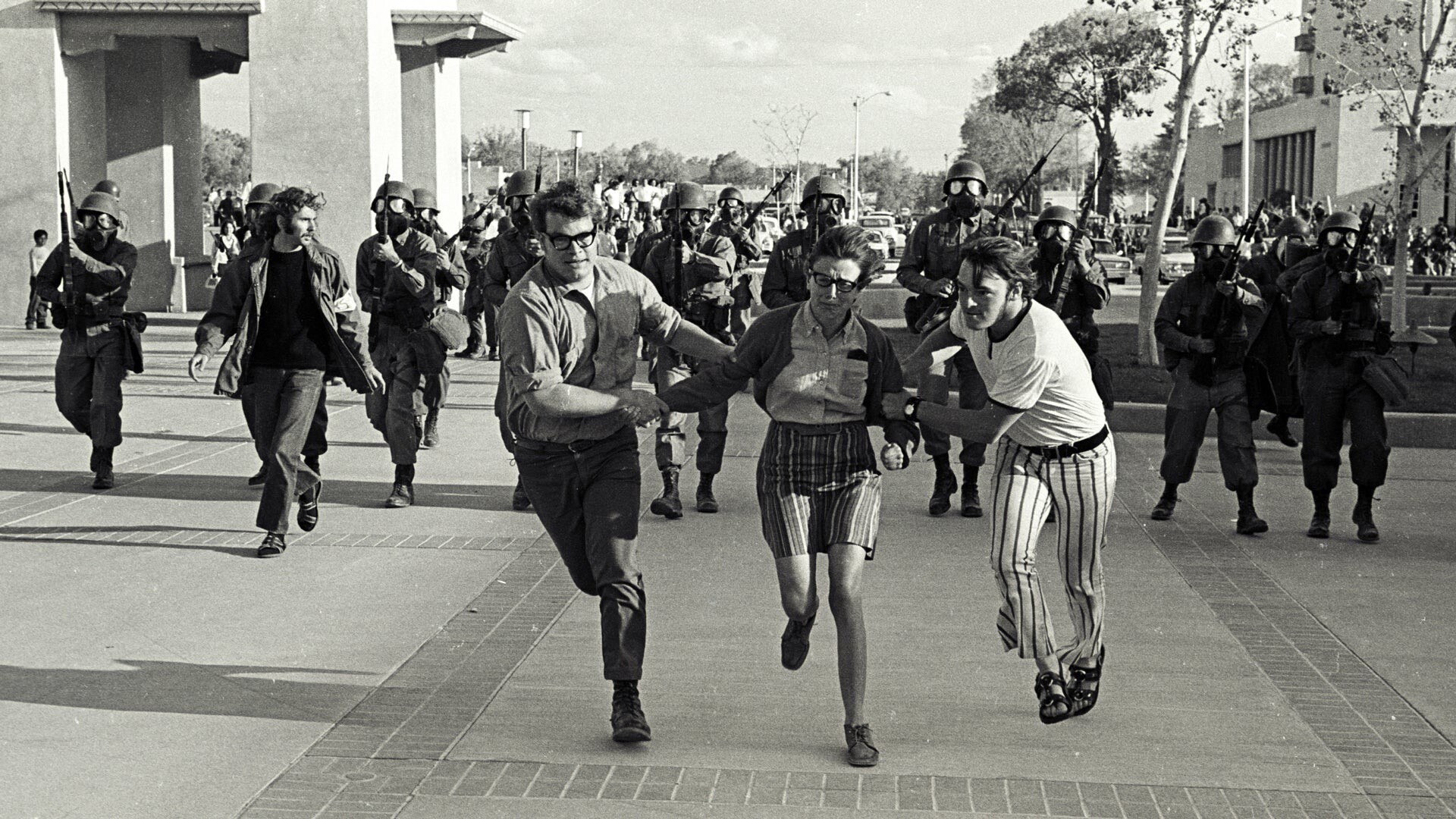 Foto em preto e branco de estudantes desarmados correndo e, ao fundo, tropas armadas da Guarda Nacional