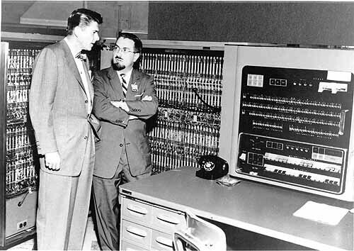 Foto em preto e branco de 2 homens de trajes sociais junto a um IBM 701, com um telefone sobre seu painel.
