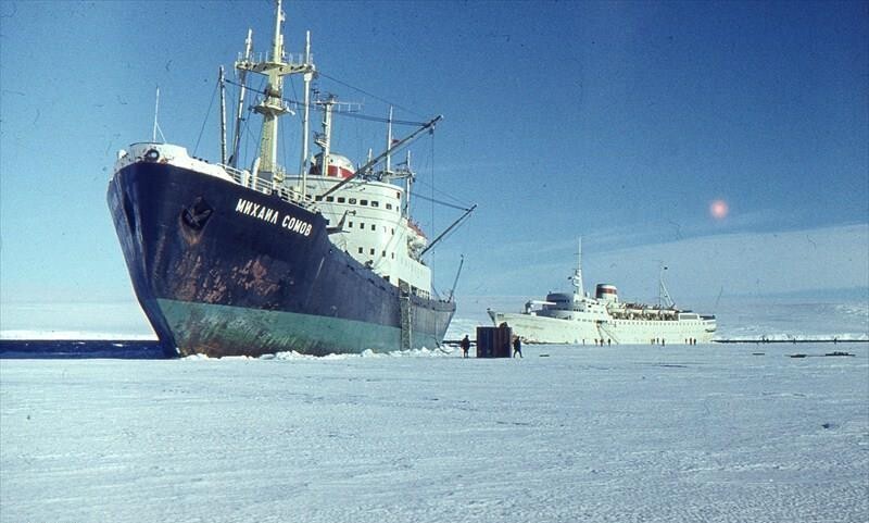 O Mikhail Somov cercado pelo mar solidificado, com pessoas em uma barraca ao redor, e um quebra-gelos de resgate ao fundo.