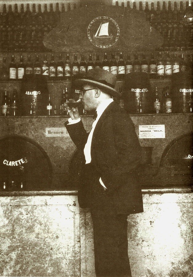 Fernando Pessoa no balcão da adega, tomando um gole.