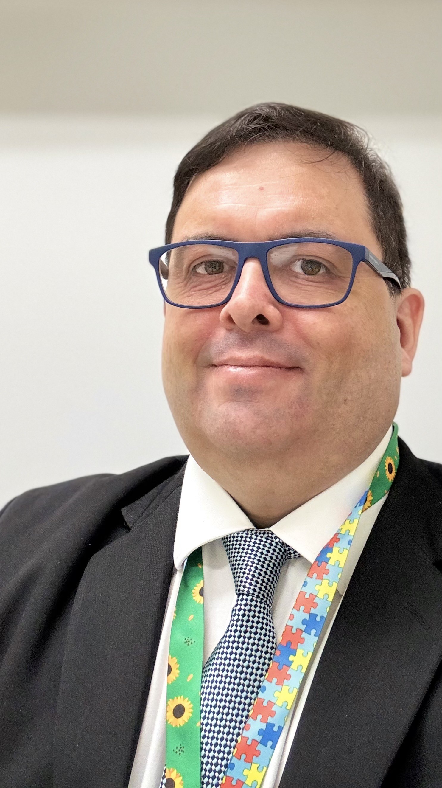 Selfie minha de terno escuro e gravata em tom azul, e com uma correia de crachá que de um lado tem o símbolo do girassol e do outro tem quebra cabeças coloridos