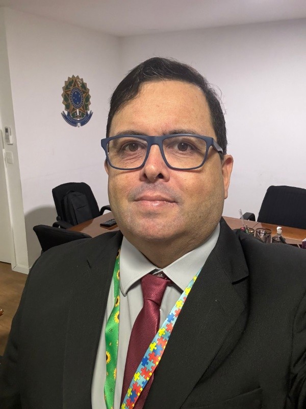 Foto minha com gravata vermelha e cordão de crachá com os símbolos do girassol e do quebra-cabeças. Ao fundo, uma mesa de reuniões e uma parede na qual pode ser visto o Brasão da República.