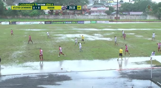 Imagem de um campo de futebol em condições precárias, com grandes banhados em vários trechos, e com os 2 times em campo.