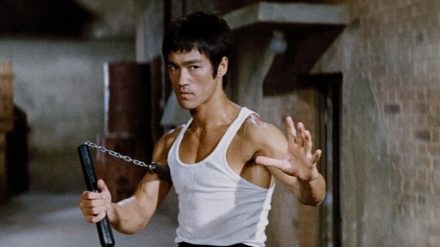 Em cena, Bruce Lee encara a câmera, segurando um nunchaku