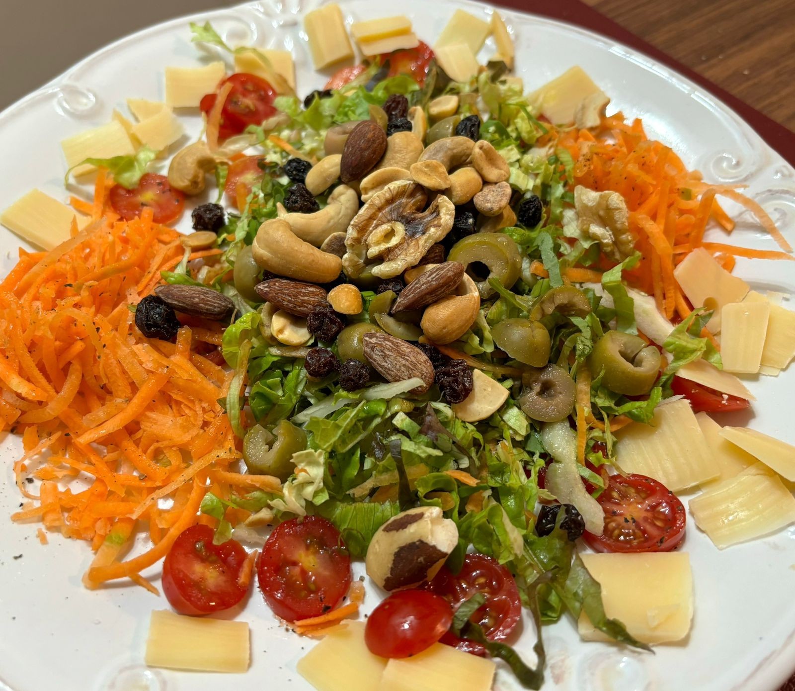 um prato de salada com folhas picadas, cenoura ralada, tomatinho em fatias, azeitona em fatias, queijo picado e castanhas diversas