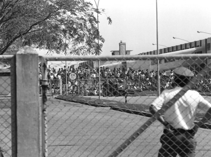 Vigilante observa os funcionários reunidos no pátio, em foto preto e branco.