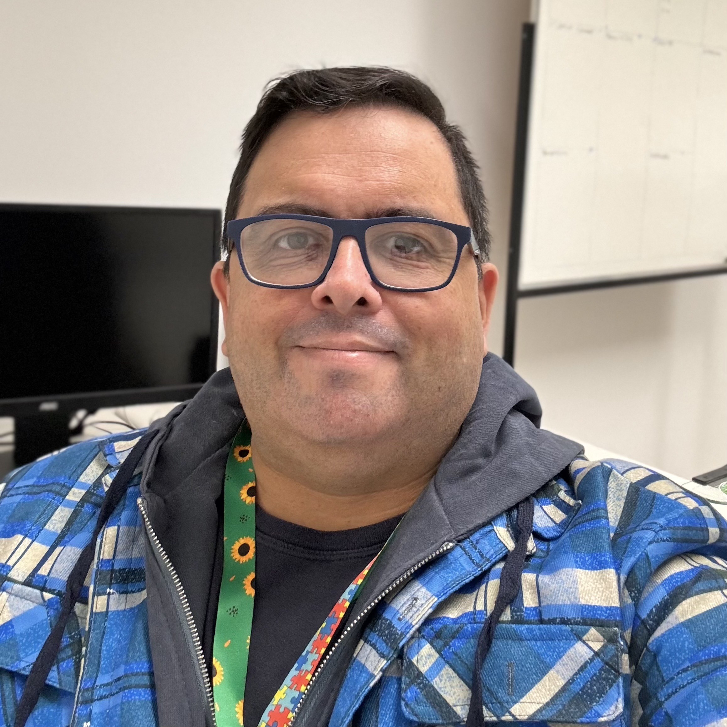 Foto minha com jaqueta em padronagem azul, camiseta azul, oculos com armação azul e correia de crachá com simbolos do autismo, em ambiente de escritório 